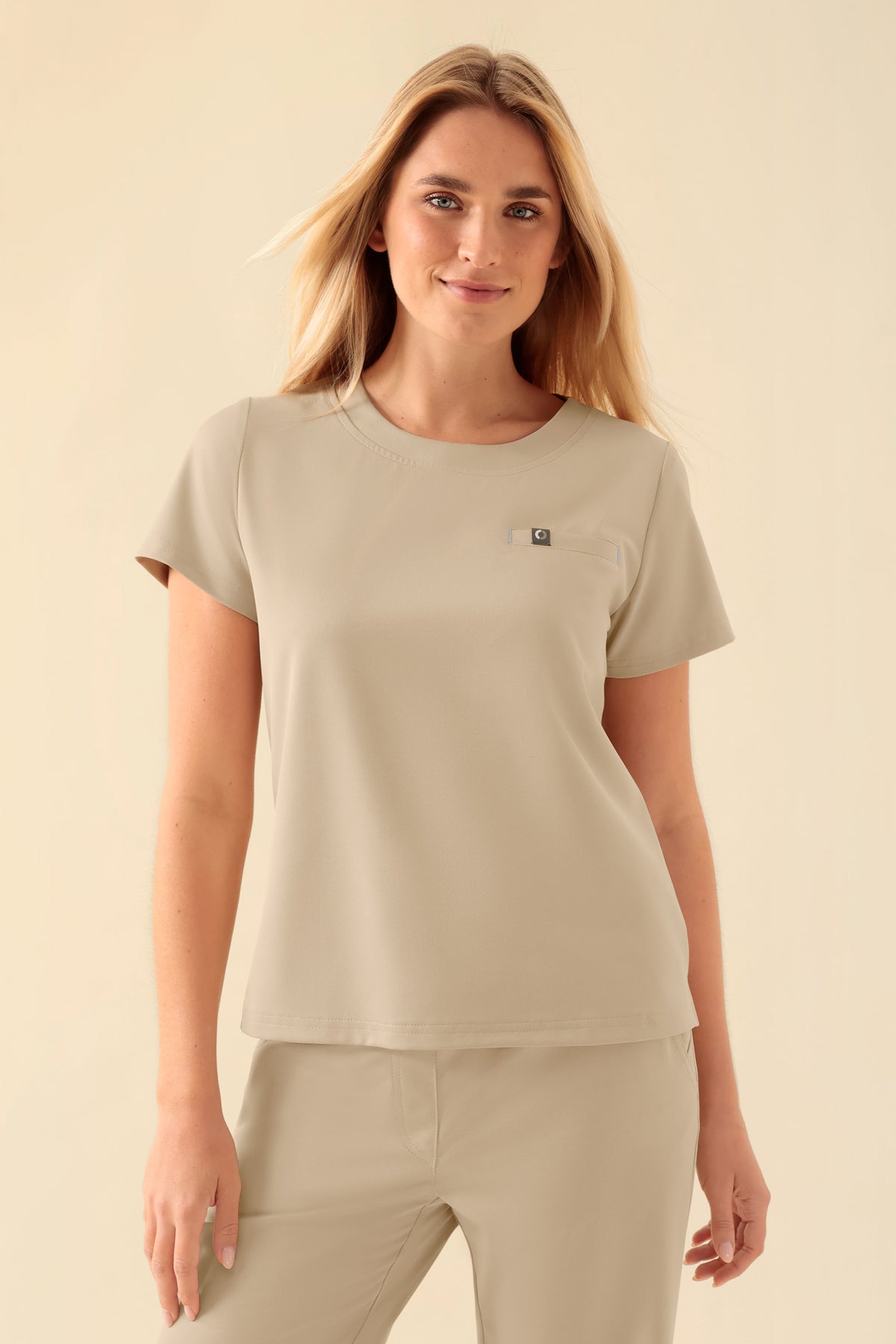 Eine Frau, die ein sandfarbenes KAERE-Shirt für Damen mit Rundhalsausschnitt, kurzen Ärmeln, Seitenschlitzen und einer kleinen Brusttasche trägt.