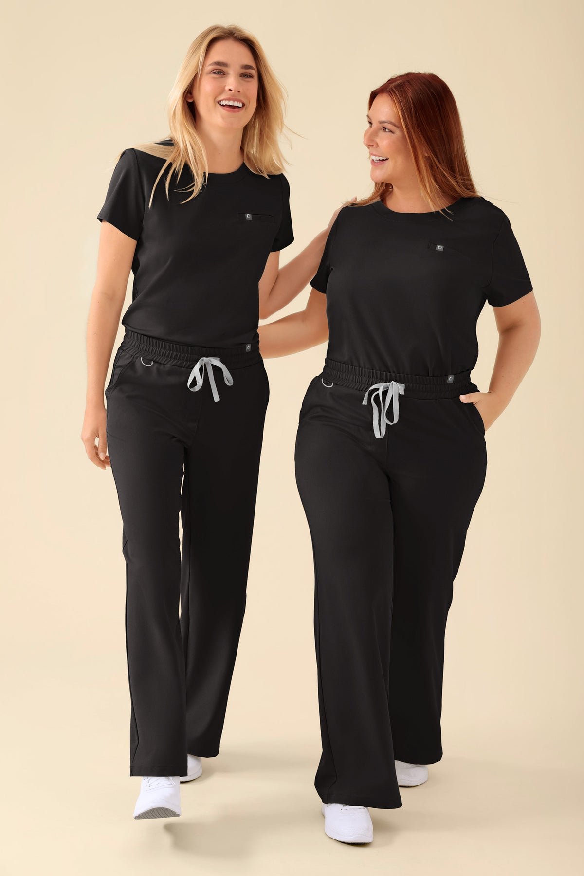 Zwei Frauen lachen, während sie identische schwarze KAERE-Shirts für Damen mit Rundhalsausschnitt und OP-Hosen mit grauen Kordelzügen tragen., Vorderansicht 