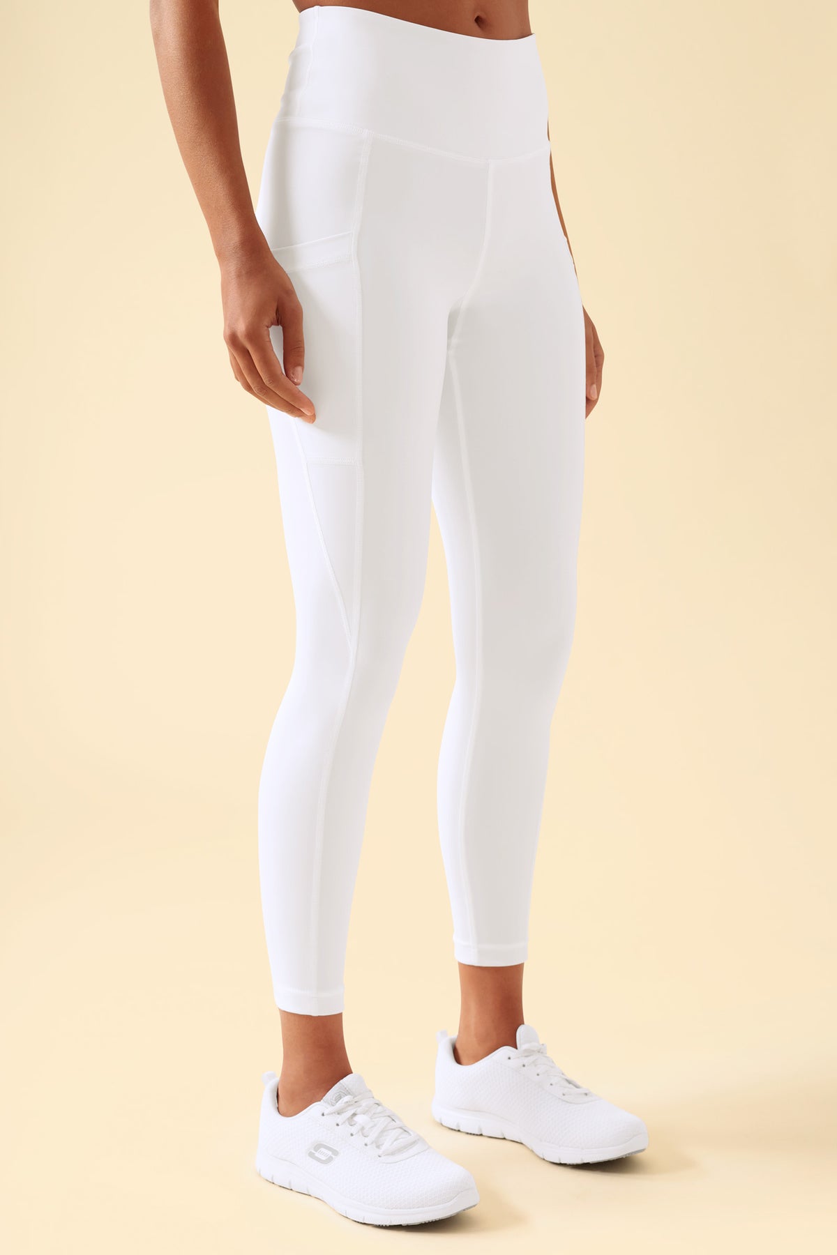 Person, die KAERE-Leggings für Damen – superelastisch, weiß – mit hoher Taille, breitem Bund, Seitentaschen und weißen Sportschuhen trägt.